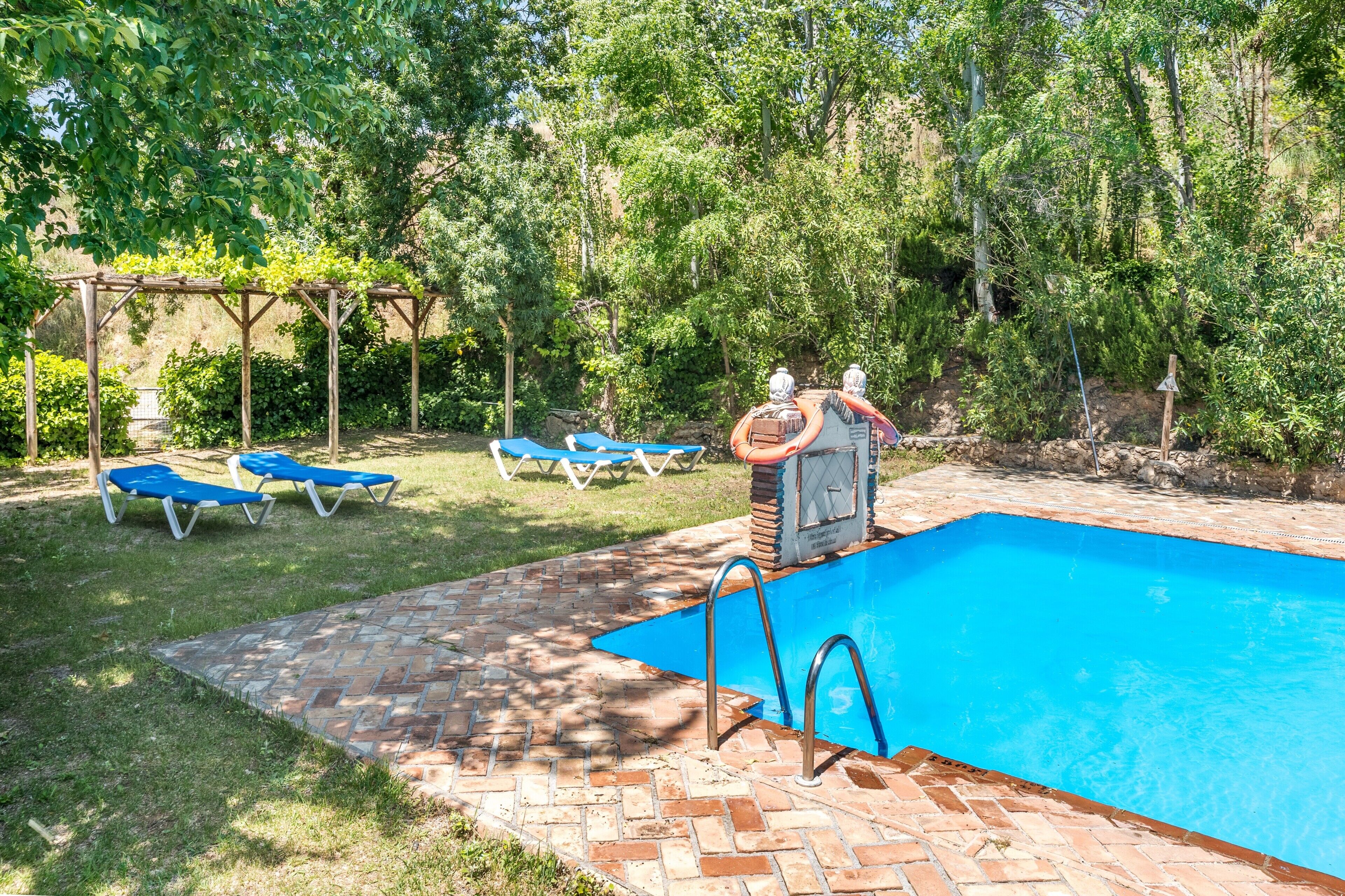 Une piscine entourée d’une terrasse en briques, de chaises longues bleues et d’une pergola couverte de plantes grimpantes.