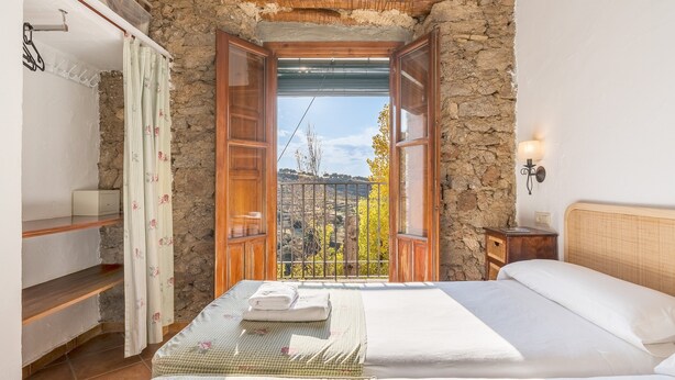 Ein Schlafzimmer mit Steinwand, einem Bett mit weißen Leinen, einem Nachttisch aus Holz und Blick durch eine offene Tür.