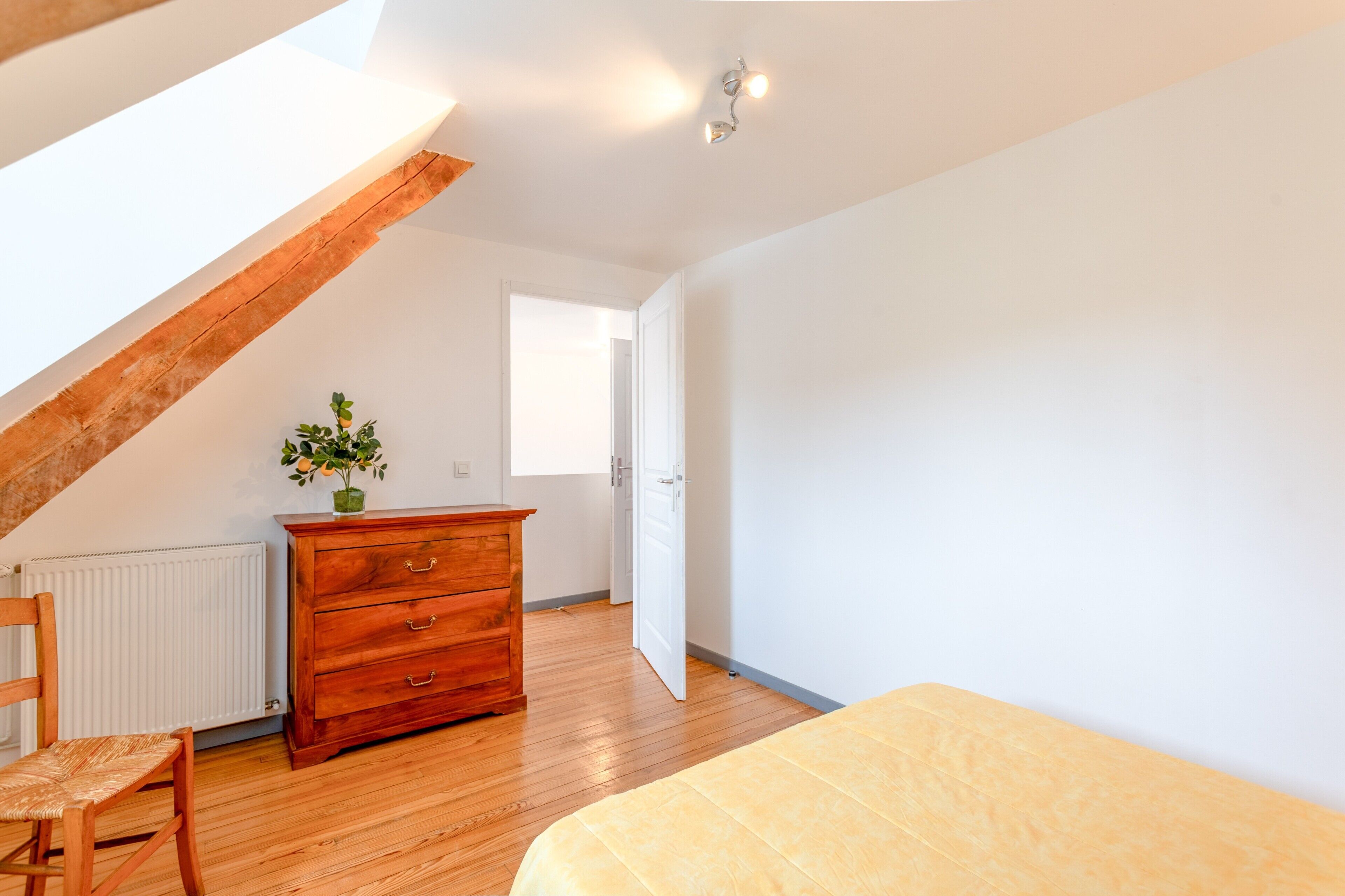 A room with a sloped ceiling, a wooden dresser, a chair, a bed, and a window.