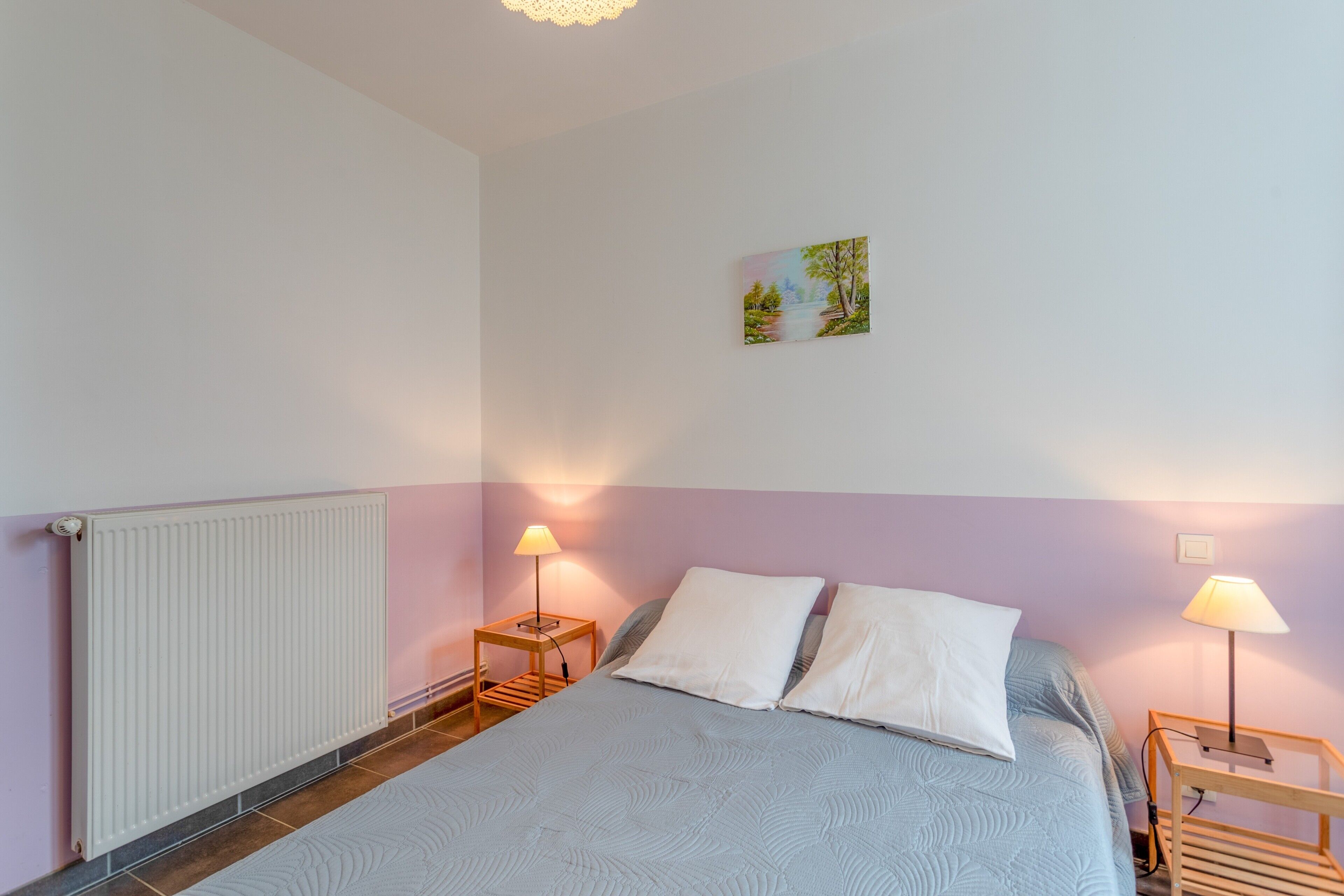 Ein Schlafzimmer mit einem Bett, zwei Nachttischlampen, einem Heizkörper und einem kleinen Gemälde an der Wand.