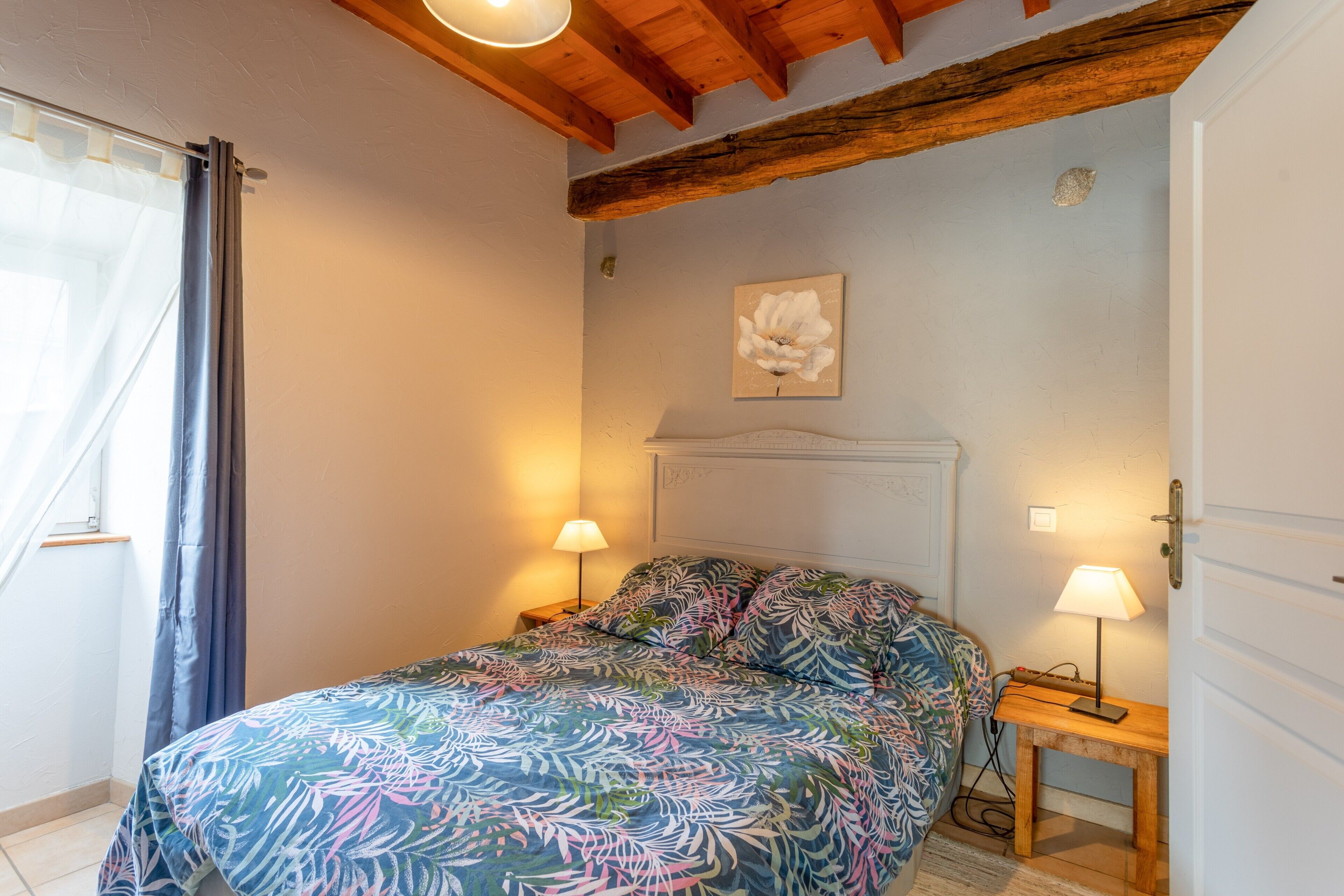 A bedroom with a bed, two bedside lamps, a wooden headboard, a wall-mounted flower painting, and a door.