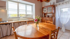 A dining area with a wooden table, chairs, a vase with flowers, a kitchen with a window, and a cabinet. - La Bergerie – Cozy House 5 min from Lourdes, Private Garden, Terrace, WiFi (Adé)