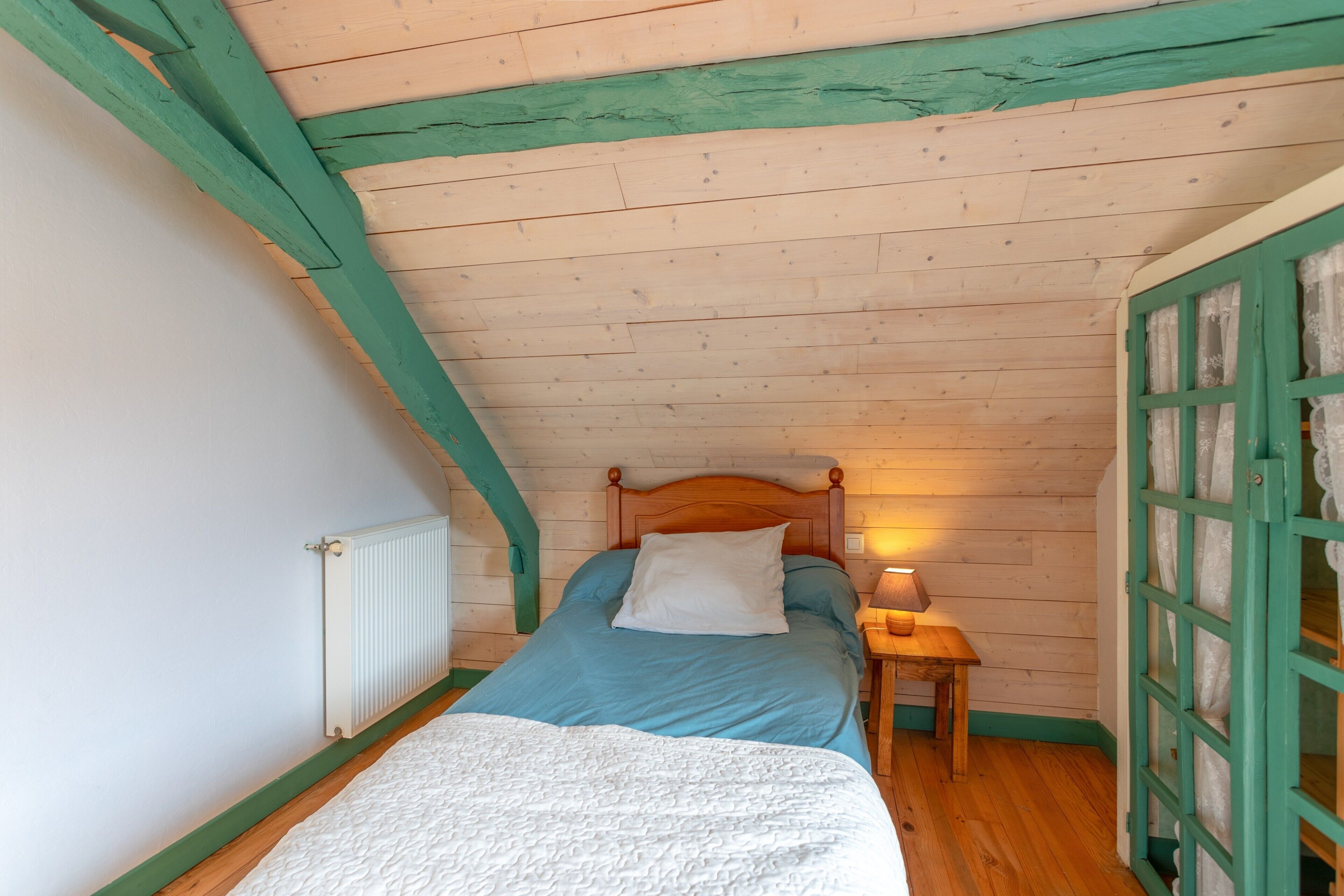 Ein Schlafzimmer mit Holzbalkendecke, einem Bett mit weißem Kopfkissen, einem Nachttisch mit Lampe und einem Fenster mit grünem Rahmen.