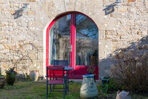 Een gebouw van steen met een deur met rode omlijsting van glas, een klein buitentafeltje met stoelen en potplanten.