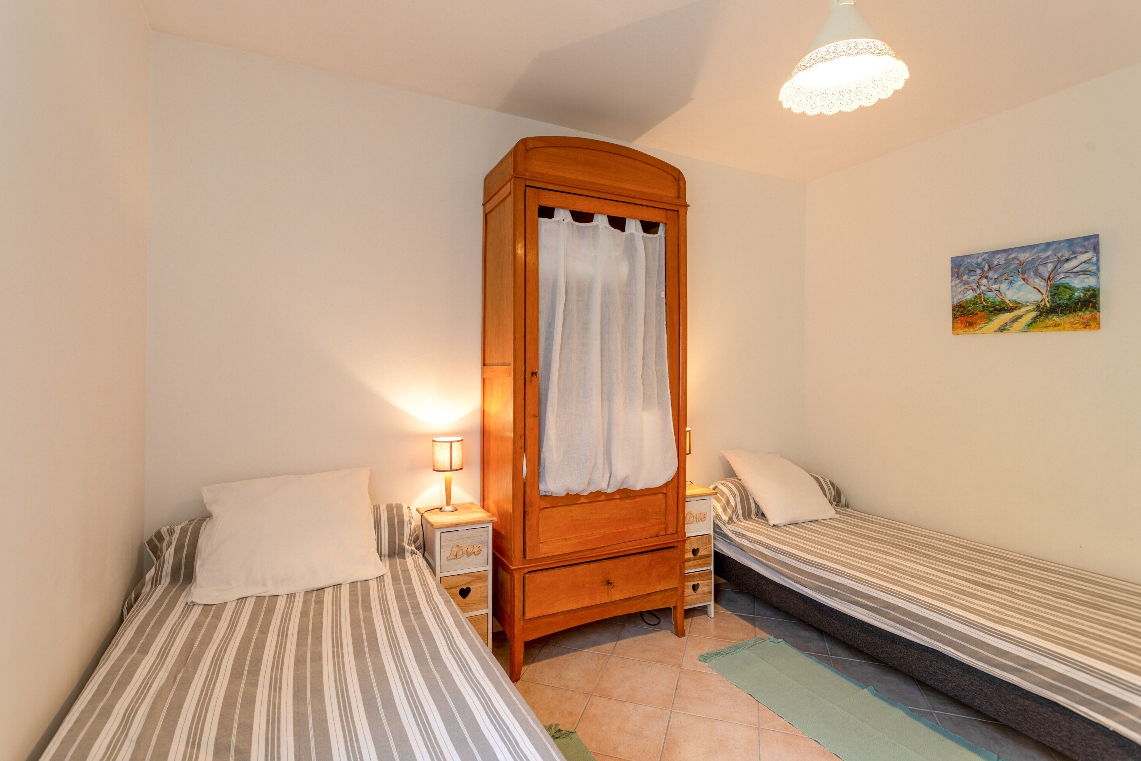 A bedroom with two single beds, a wooden wardrobe, a nightstand with a lamp, and a painting on the wall.
