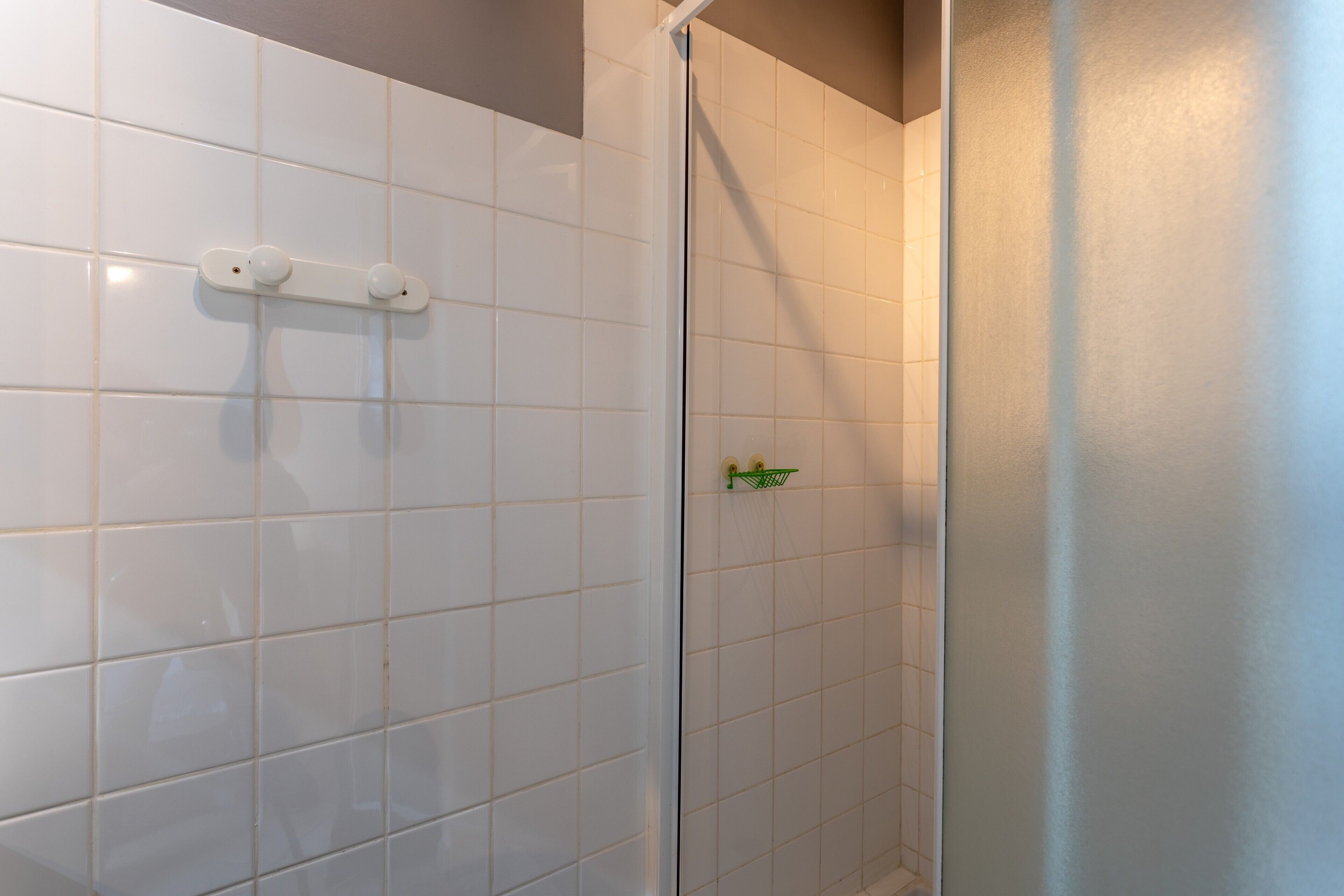 Ett badrum med vita kakelväggar, en dusch med ett grönt handtag och en vit handdukshängare.