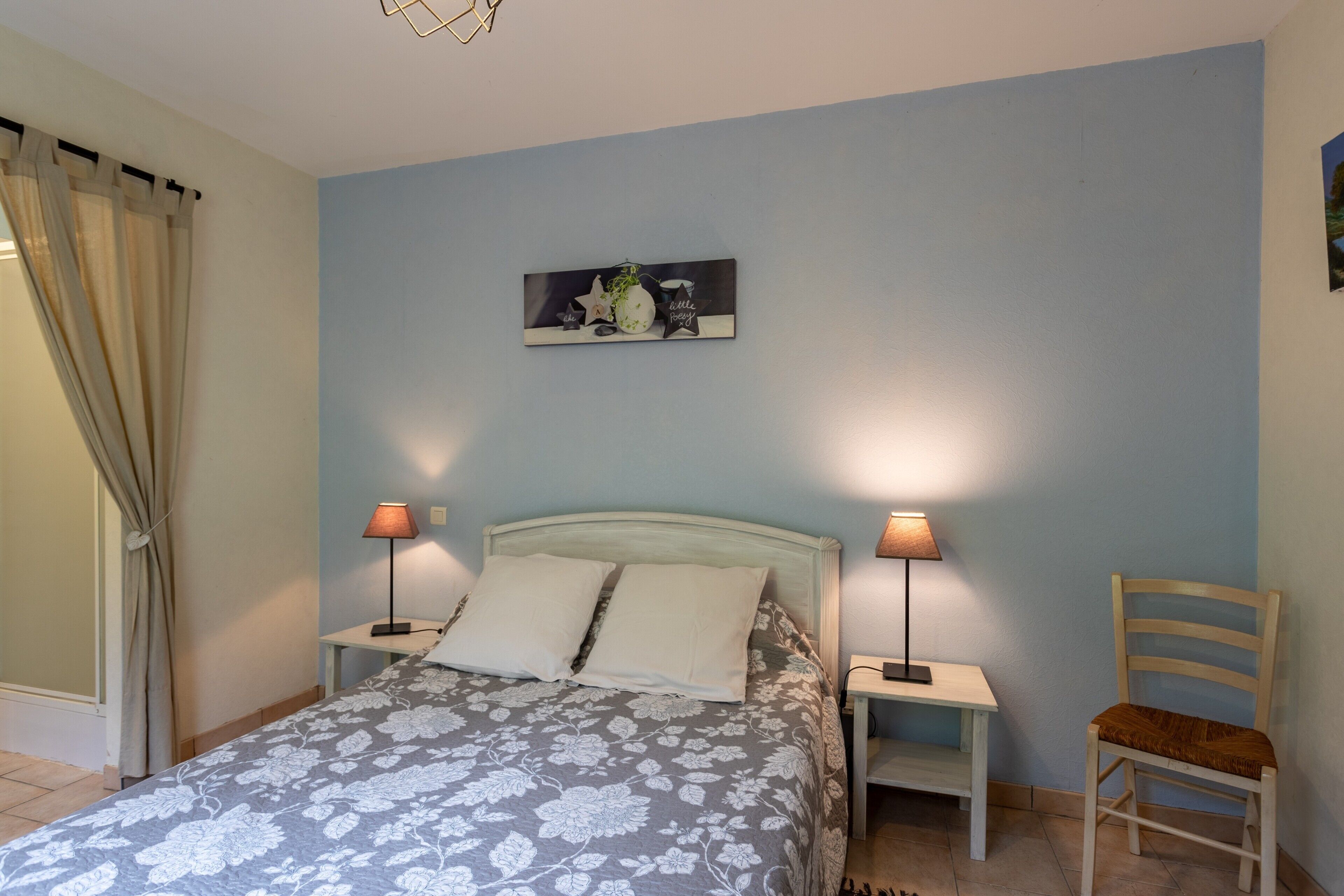 A bedroom with a bed, two bedside lamps, a chair, a wall-mounted picture, and a curtain.