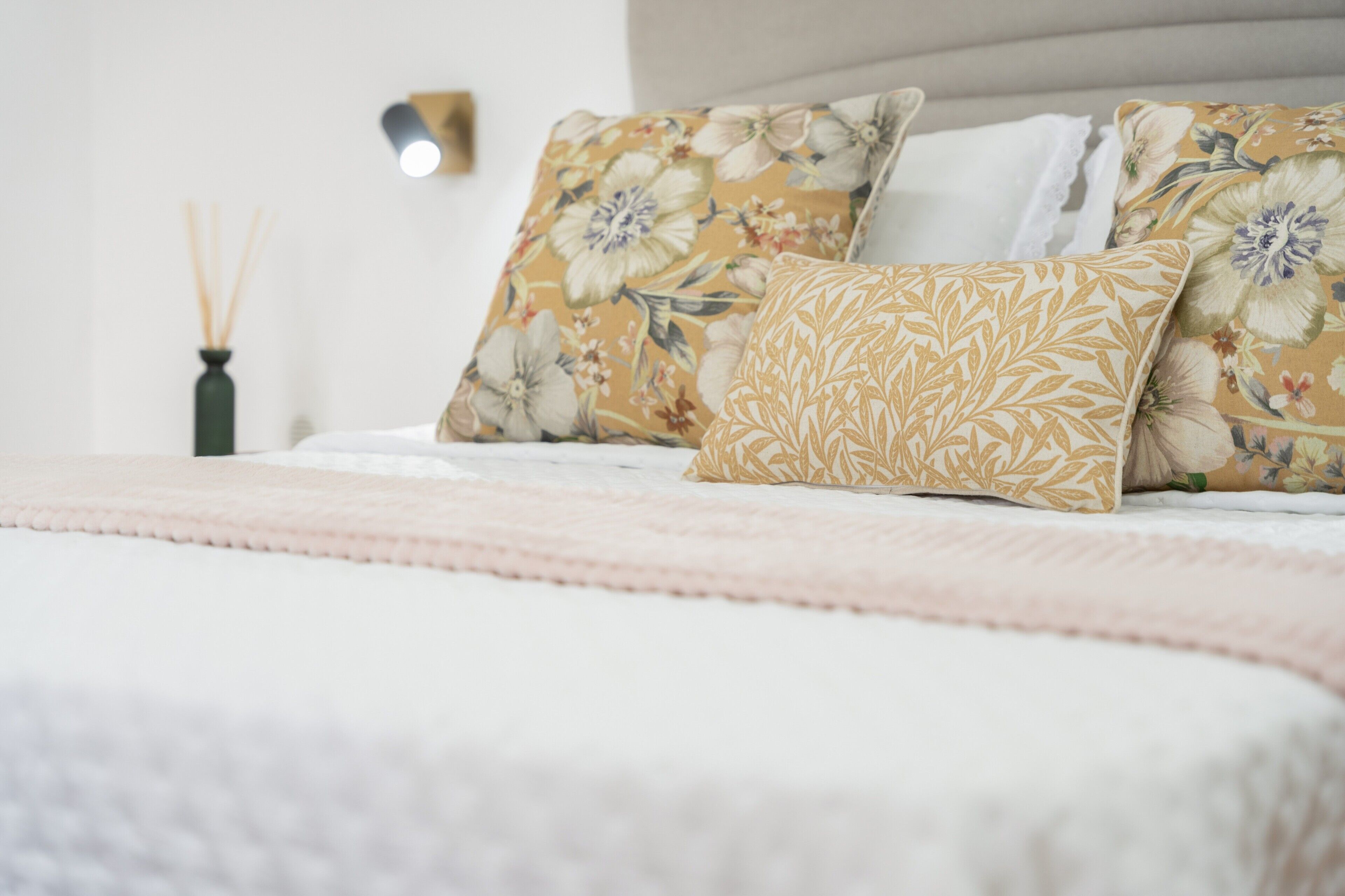 Ein ordentlich bezogenes Bett mit blumigem Kissenmuster, ein Nachttisch mit einer Vase und eine Wandleuchte.