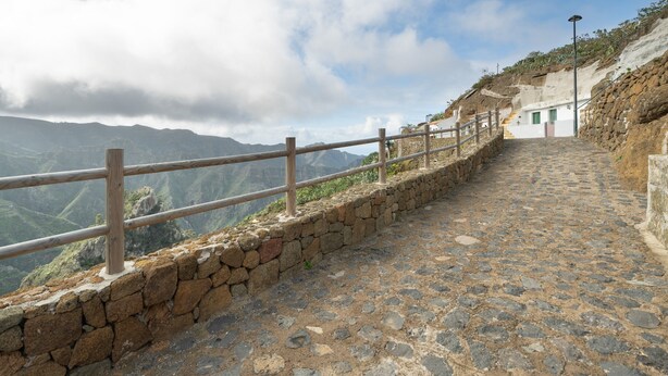 A stone pathway leading to a building with a wooden railing, surrounded by mountains and a cloudy sky.