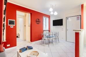 A room with a red wall, a dining table with chairs, a TV mounted on the wall, and a door with a 'No Entry' sign.
