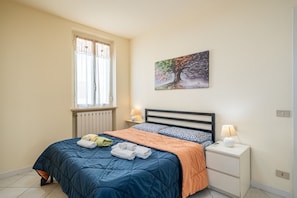 A bedroom with a bed, a nightstand, a lamp, a window with curtains, and a painting on the wall.