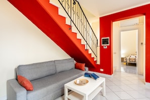 A living room with a grey sofa, a white coffee table with books, a red staircase, and a doorway leading to another room.