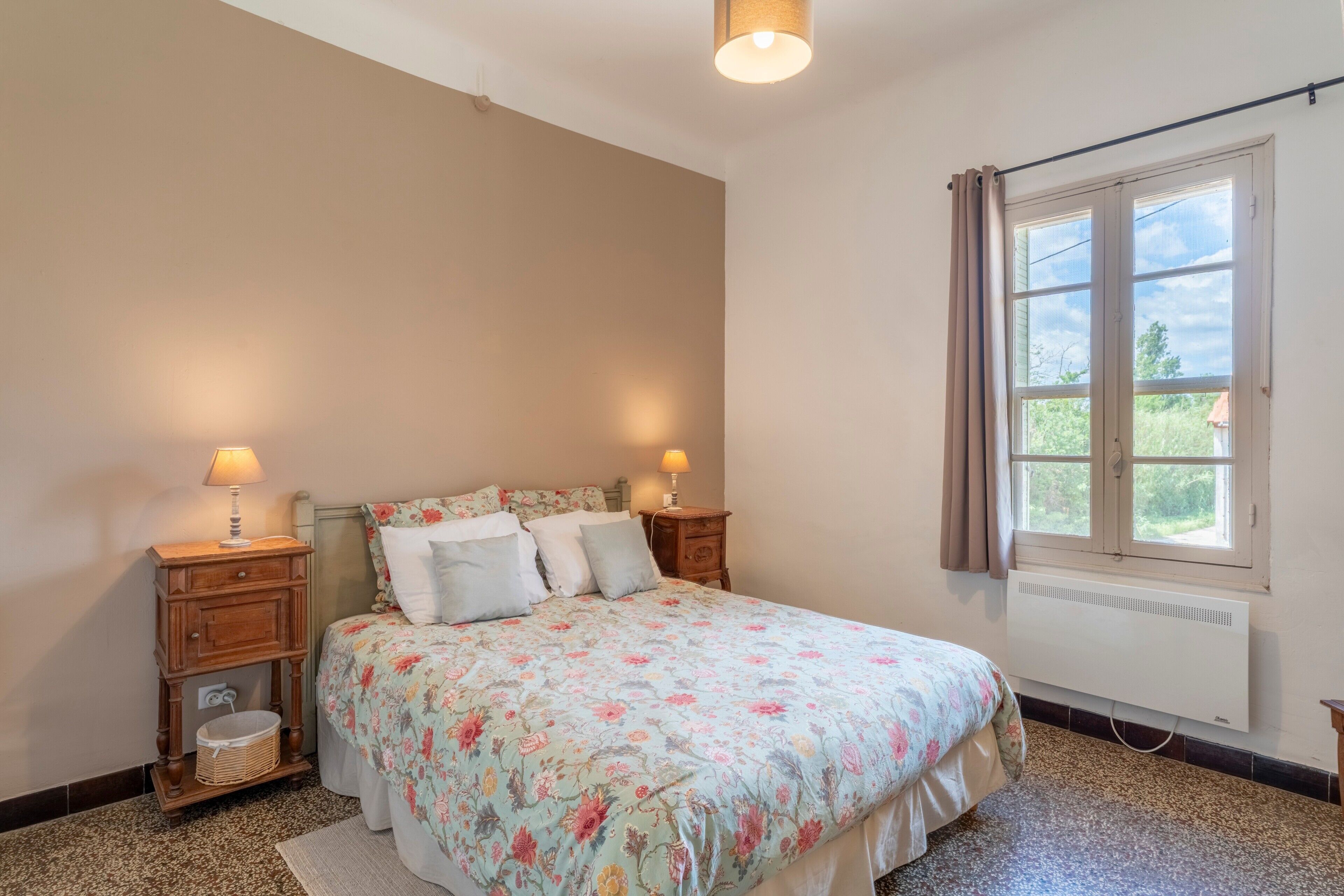Une chambre à coucher avec un lit, deux lampes de chevet, une table de chevet, un radiateur et une fenêtre avec des rideaux.