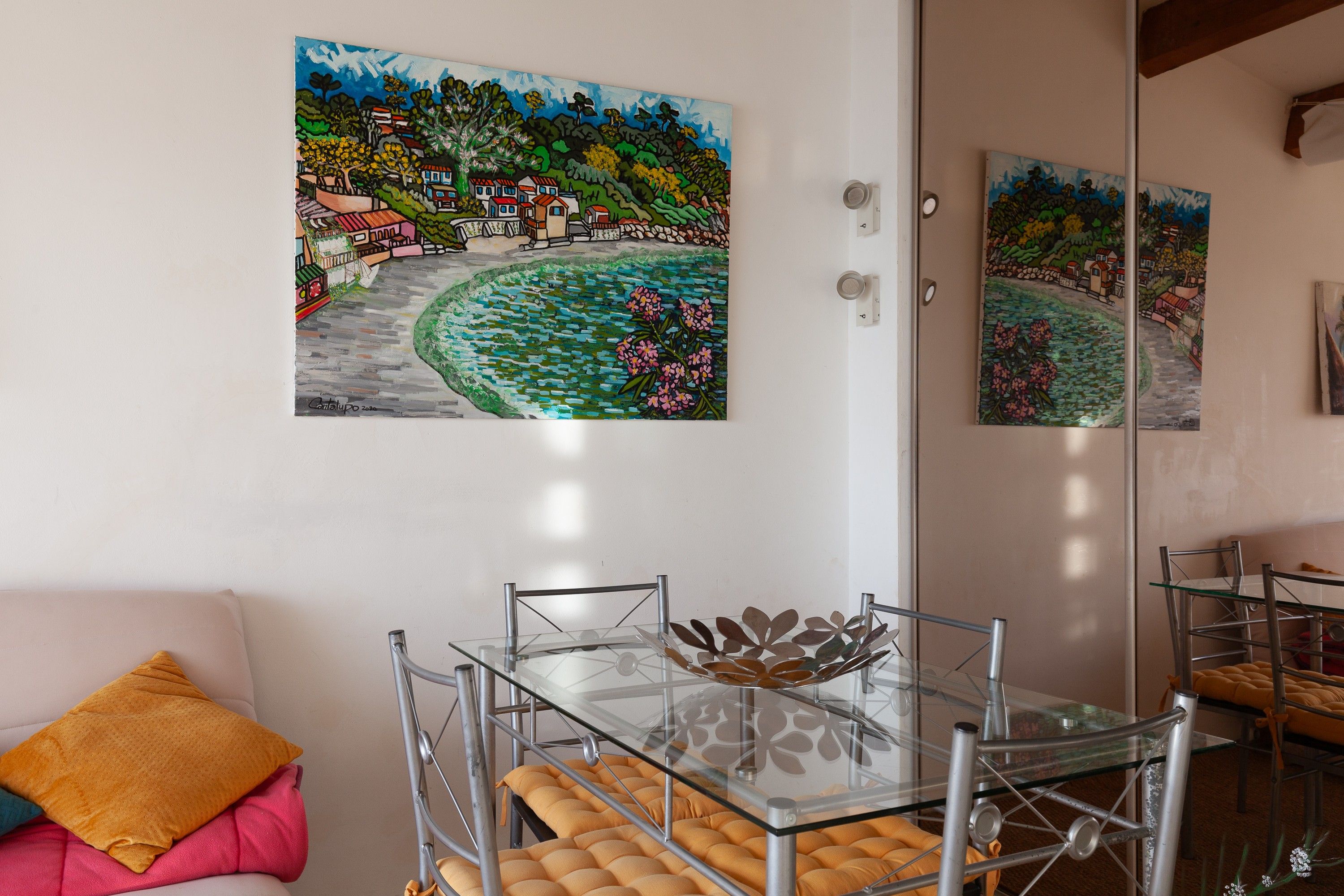 A dining area with a glass table and metal chairs, a sofa with cushions, and two paintings on the wall.
