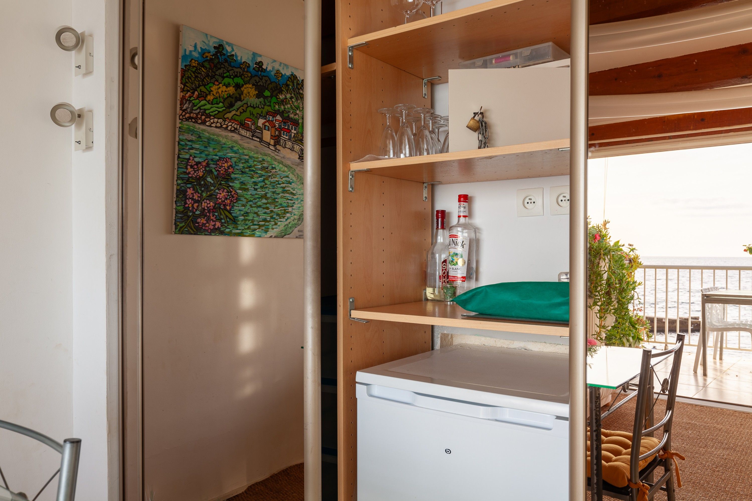 Ein Zimmer mit einem weißen Minikühlschrank, einem Regal mit Gläsern und einer Flasche, einem Bild an der Wand und Blick aufs Meer.