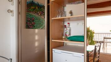 Ein Zimmer mit einem weißen Minikühlschrank, einem Regal mit Gläsern und einer Flasche, einem Bild an der Wand und Blick aufs Meer.