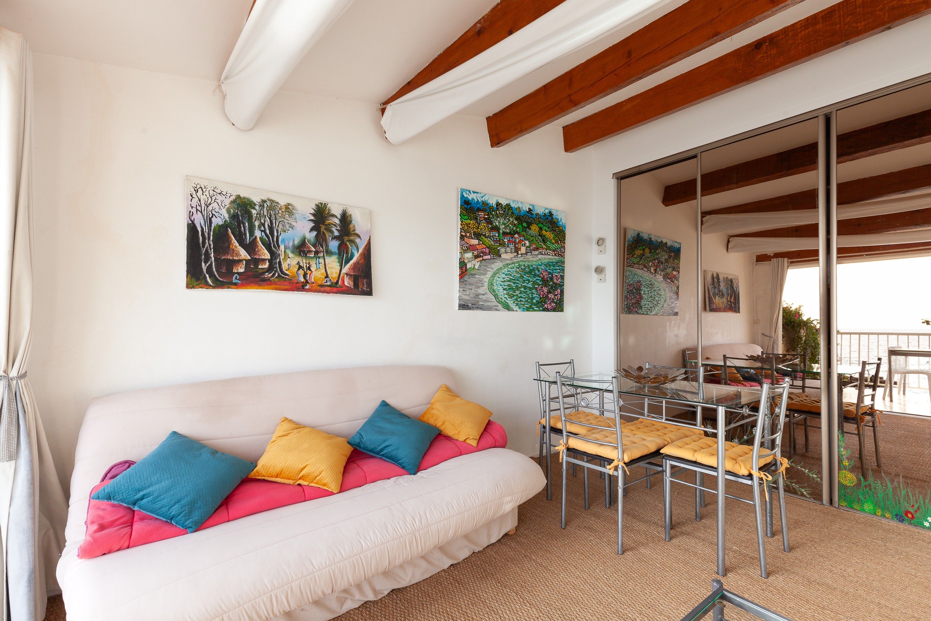 A room with a sofa, cushions, a glass table, chairs, and artwork on the walls.