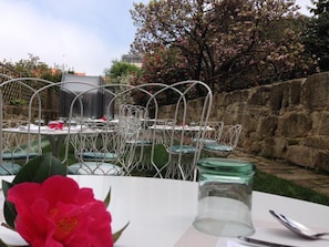 Un espace extérieur aménagé pour les repas, avec des chaises et des tables en métal, un mur en pierre et un arbre en fleurs.