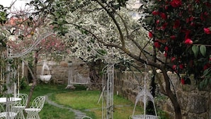 Un jardín con sillas y mesas de metal blanco, un muro de piedra y árboles en flor.