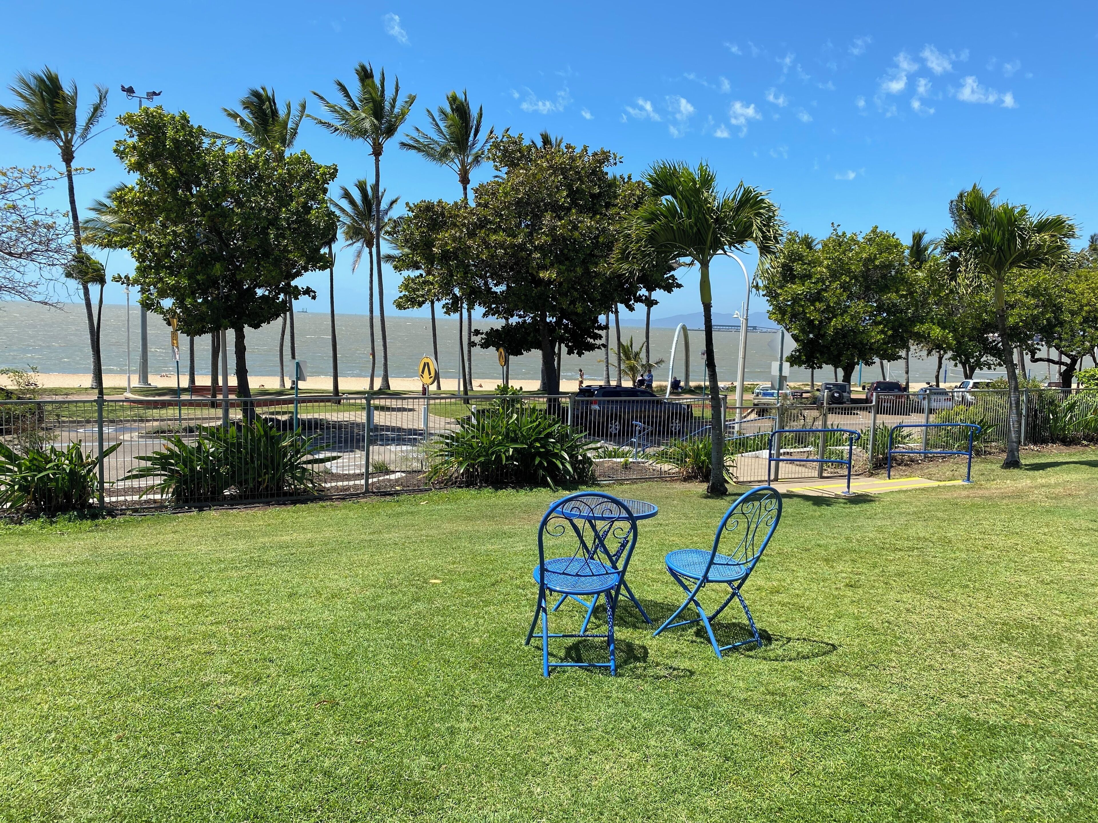 A grassy area with blue metal chairs and a table, palm trees, and a view of the sea.