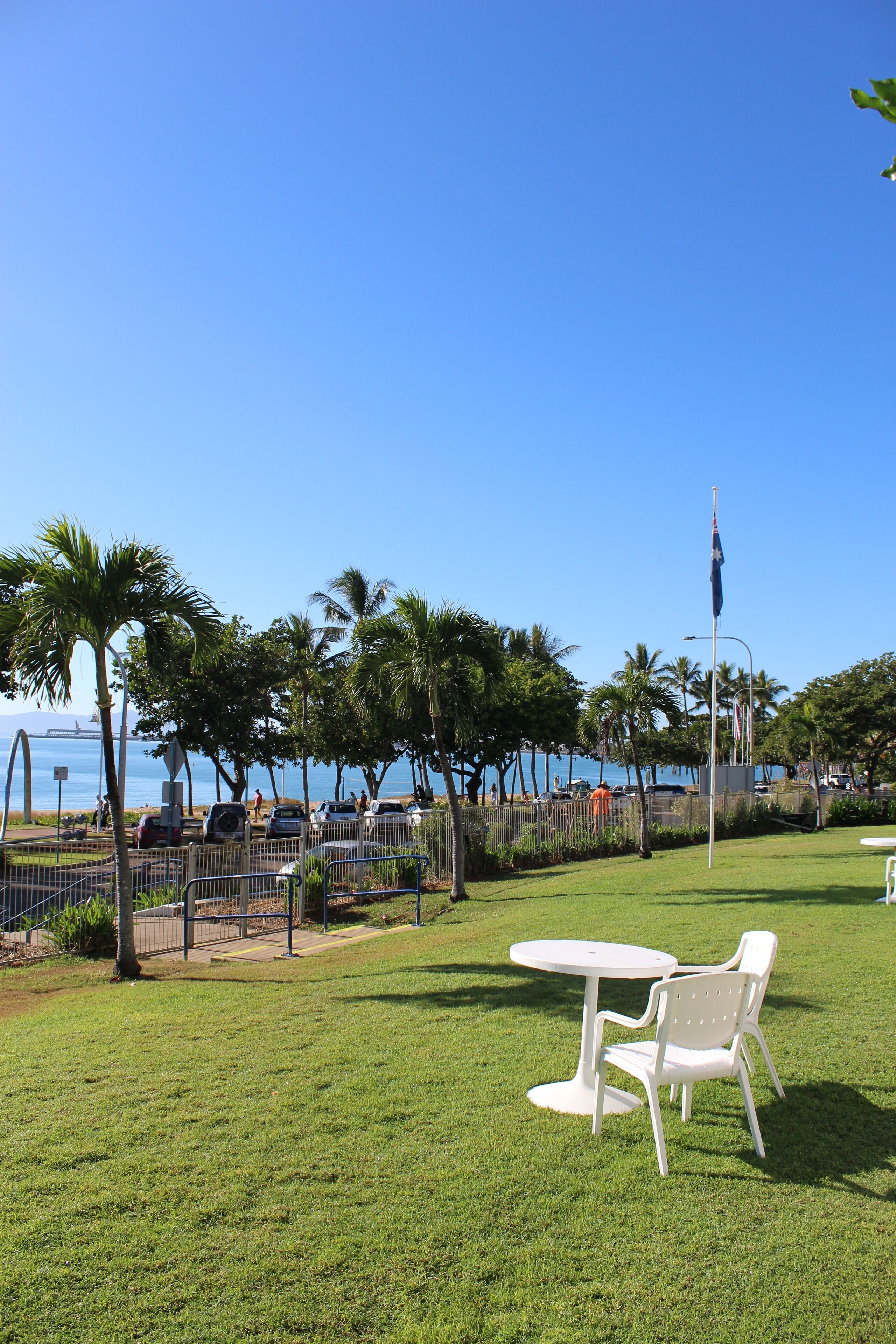 A grassy outdoor area with a white table and chair, palm trees, and a view of the ocean.
