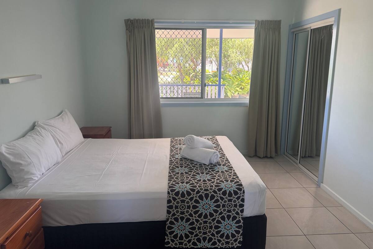 A bedroom with a bed, a bedside table, a window with curtains, and a view of greenery outside.