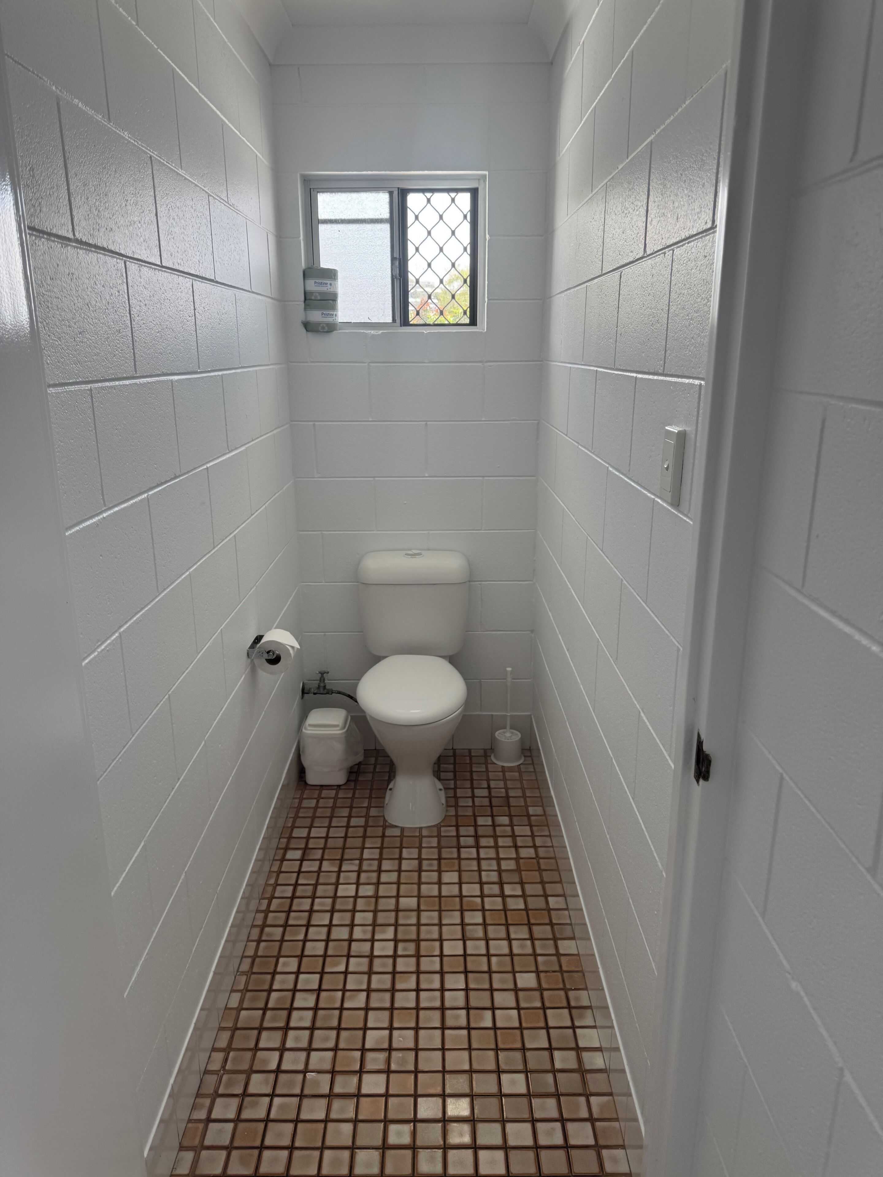 A small, narrow bathroom with white tiled walls and floor, a toilet, a small trash bin, and a window with a metal grid.