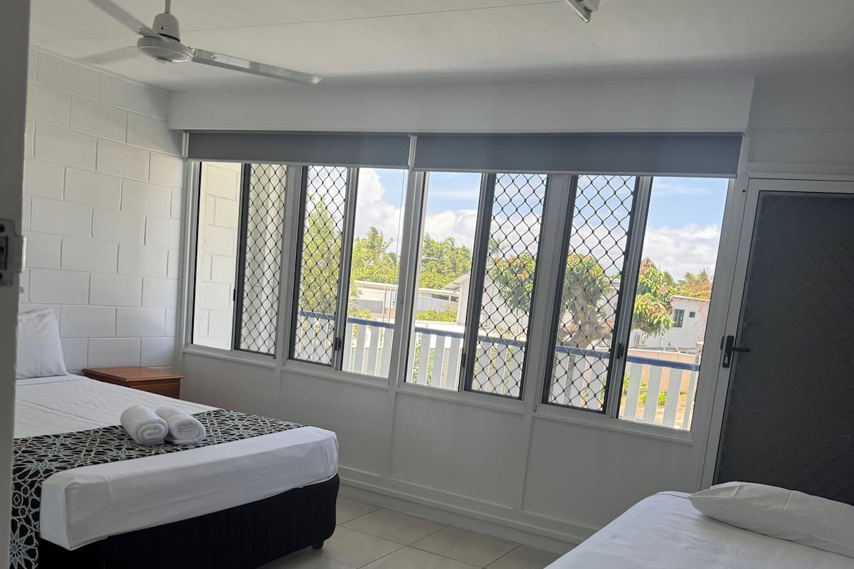 A hotel room with two beds, a ceiling fan, and a large window with a view of the neighborhood.
