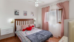 Ein Schlafzimmer mit einem Bett, Nachttischen, einem Deckenventilator, einem Fenster mit Vorhängen und Wandbildern.