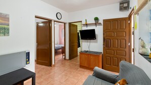 Ein Zimmer mit einer Couch, einem an der Wand montierten Fernseher, einer Holztür und einem gefliesten Boden.