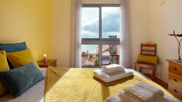 Une chambre d’hôtel comprenant un lit, une table de chevet, une chaise, une commode et une fenêtre donnant sur des immeubles.
