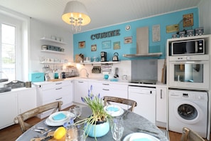 Una cocina con armarios blancos, pared azul y una mesa de comedor dispuesta con platos, cubiertos y un florero con flores.