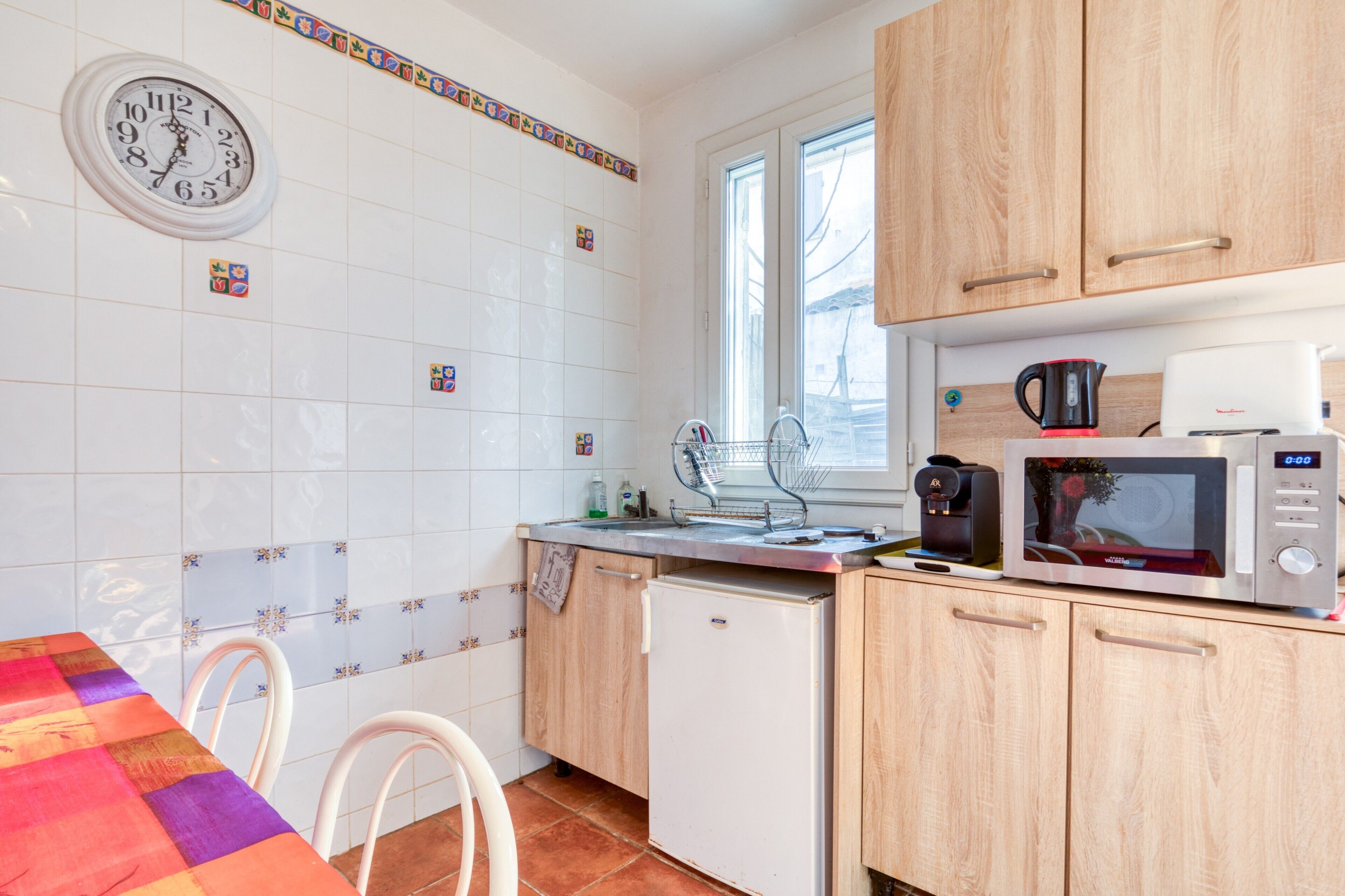 Una cocina con baldosas blancas, un reloj, armarios de madera, un refrigerador, un microondas y una ventana.