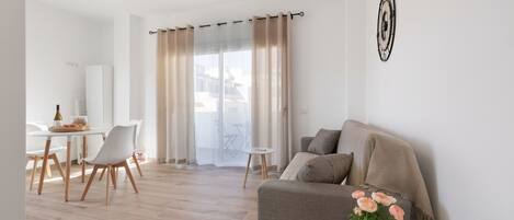 A modern living room with a sofa, a small round table with a plant, a dining area with chairs, and a view through a large window.