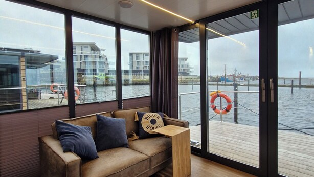 A modern living room with a sofa, a wooden side table, and a view of the water through large windows.