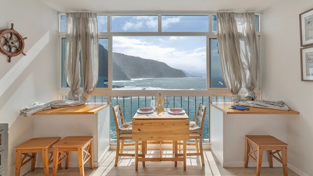 Ein Essbereich mit Tisch und Stühlen, einer hölzernen Bank, einer Schiffslenken-Dekoration und Blick auf das Meer durch ein großes Fenster.