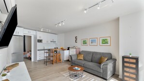 Eine moderne Küche mit weißen Schränken, ein Essbereich mit Hockern, ein graues Sofa und ein kleiner Couchtisch.