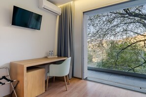 Une chambre moderne dotée d’un bureau en bois, d’une chaise, d’un téléviseur à écran plat et d’une grande fenêtre donnant sur les arbres.