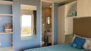 A bedroom with a bed, a mirror, a bathroom, and a shelf with plants. - Mobile home with terrace with lake view, pool and air conditioning (Padenghe sul Garda)