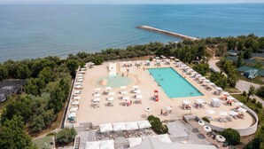 Vista aérea de un complejo turístico con una gran piscina, zona de playa y un muelle que se adentra en el mar.