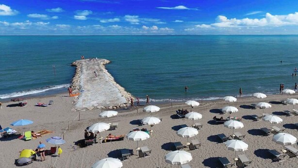 Una playa con numerosas sombrillas blancas y tumbonas, un espigón rocoso y un cielo azul despejado.