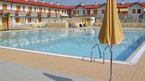 Une piscine avec un parasol jaune, une terrasse carrelée et, au fond, des bâtiments aux toits rouges.