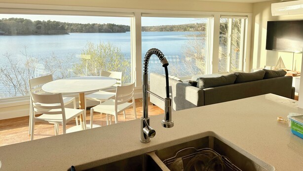 Een moderne keuken met een groot raam dat uitkijkt over een meer, een zithoek met stoelen, en een woonkamer met een bank.