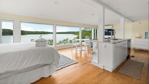 Een slaapkamer met een groot raam met uitzicht op een meer, een bed met witte lakens, een kleine kitchenette en een uitzicht op de natuur.