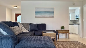 A living room with a dark blue sofa, a small side table, a wicker chair, and a framed landscape painting on the wall.