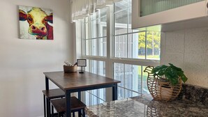A room with a dining table, chairs, a painting, a window with blinds, and a plant.