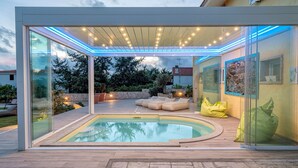 Un espace piscine extérieur moderne, doté de murs en verre, d’un espace salon couvert et d’un chemin en pierre.