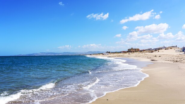 A beach with clear blue water, sandy shore, and a few buildings in the distance. - Ammentos by Interhome (Marina di Sorso)