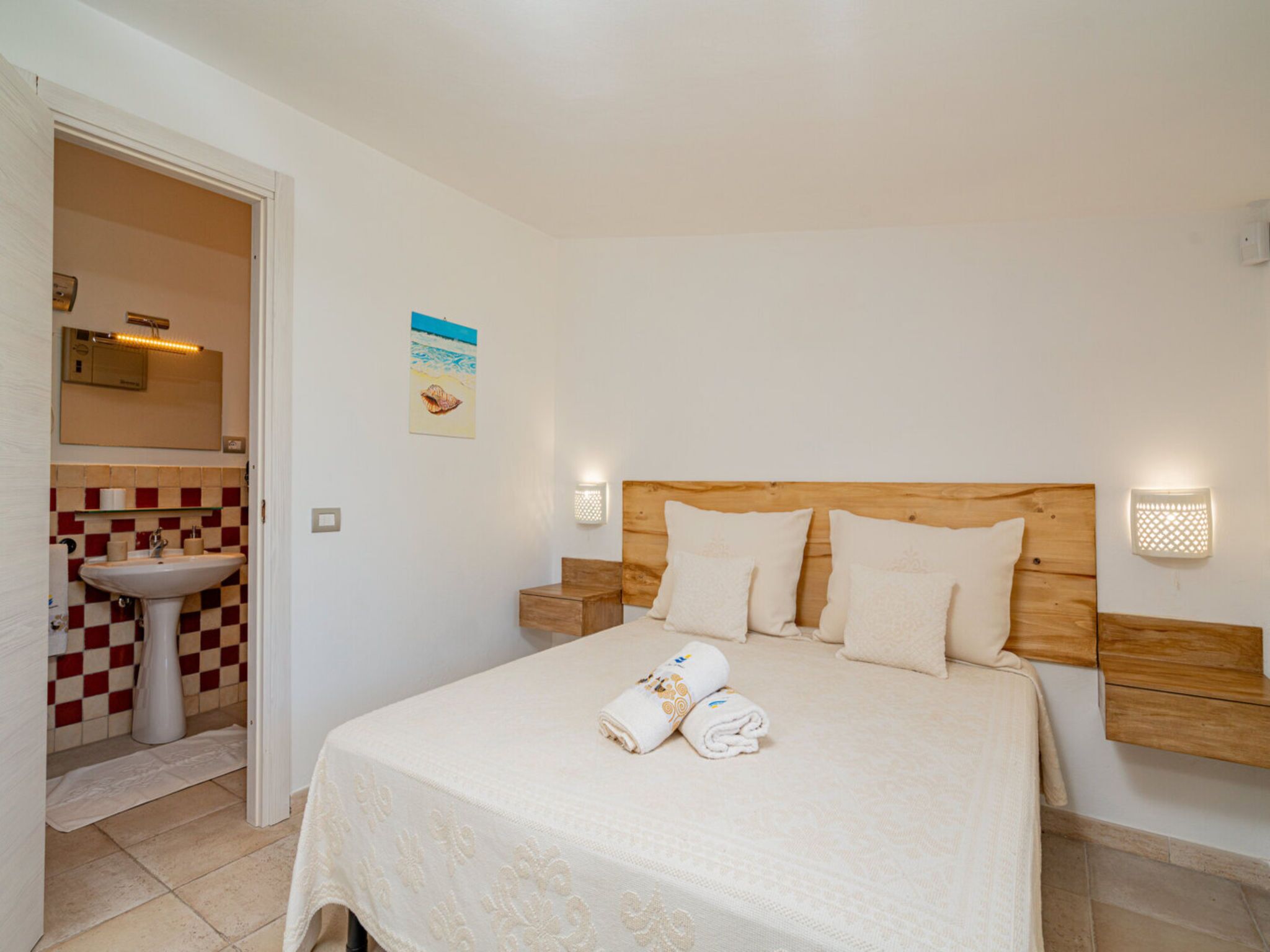 A hotel room with a bed, a wooden headboard, two bedside tables, a bathroom with a sink and mirror, and a checkered tile backsplash.