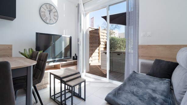 Un salon moderne doté d’un téléviseur à écran plat, d’une horloge, d’un coin repas avec une table et des chaises, et d’une porte-fenêtre coulissante donnant sur une terrasse extérieure.